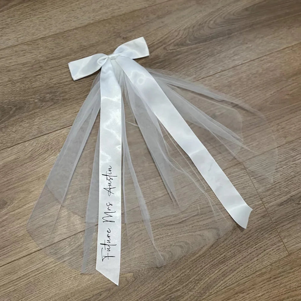 Personalized 'Future Mrs' bow veil in white tulle with satin bow, shown on bachelorette bride accessory display.