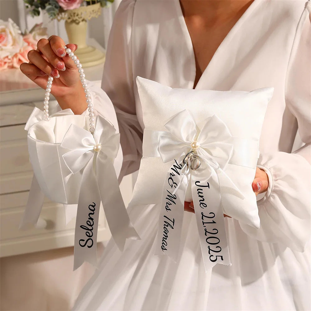 Personalized white flower girl basket and ring bearer pillow set with custom lettering, shown on wedding ceremony display.