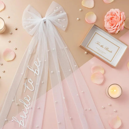 White pearl hair bow clip with attached tulle veil, shown on bride-to-be bachelorette accessory display.