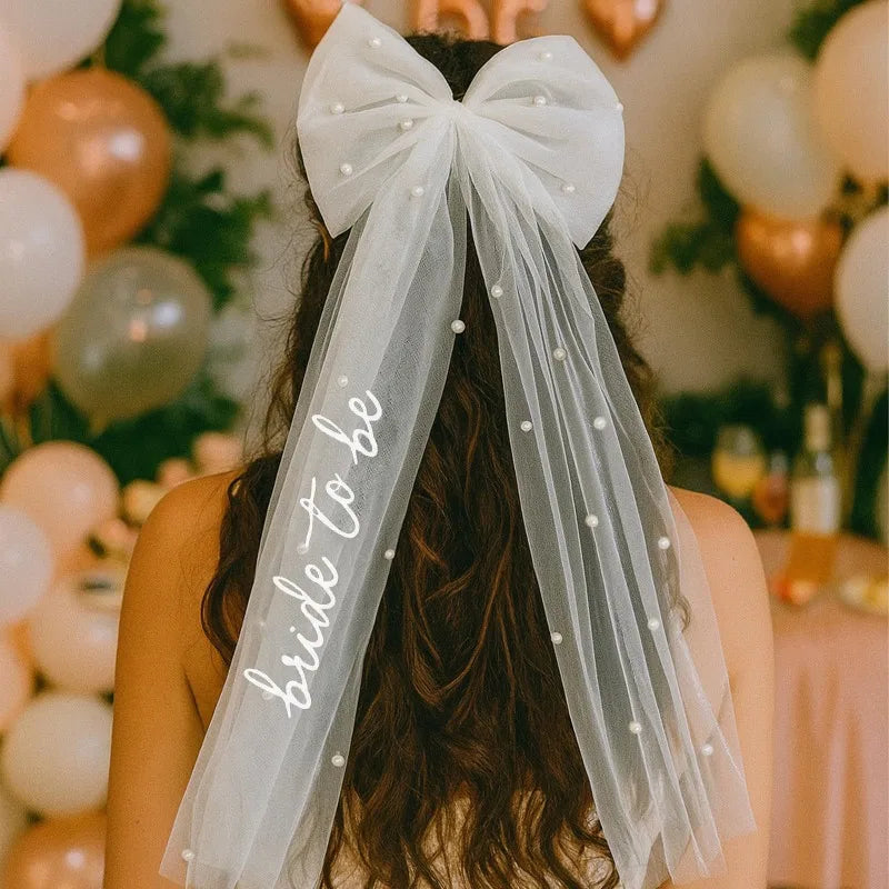 White pearl hair bow clip with attached tulle veil, shown on bride-to-be bachelorette accessory display.