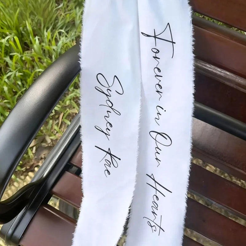 Personalized white memorial chair ribbon with 'Forever In Our Hearts' printed lettering, shown on reserved wedding ceremony chair.