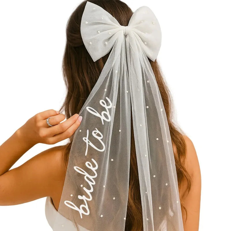 White pearl hair bow clip with attached tulle veil, shown on bride-to-be bachelorette accessory display.