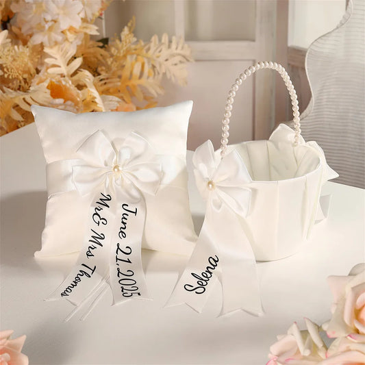 Personalized white flower girl basket and ring bearer pillow set with custom lettering, shown on wedding ceremony display.
