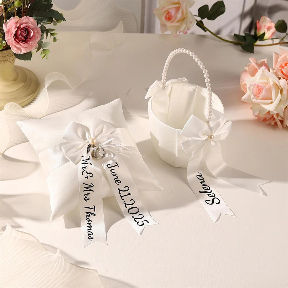 Personalized white flower girl basket and ring bearer pillow set with custom lettering, shown on wedding ceremony display.