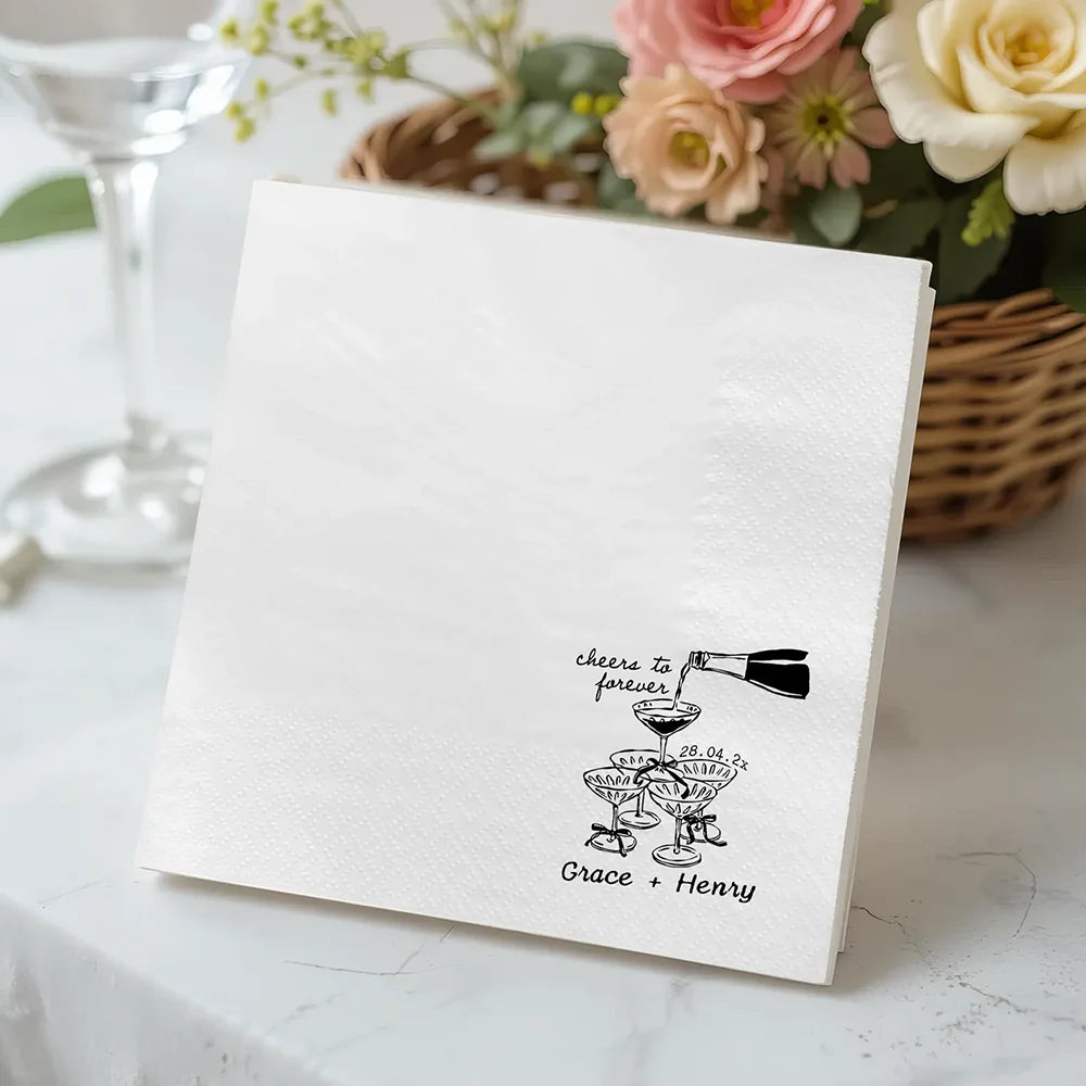 Custom 'Cheers To Forever' cocktail napkins personalized with names and wedding date, shown on champagne tower wedding reception display.