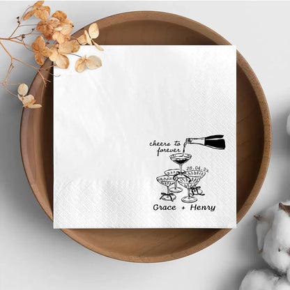 Custom 'Cheers To Forever' cocktail napkins personalized with names and wedding date, shown on champagne tower wedding reception display.