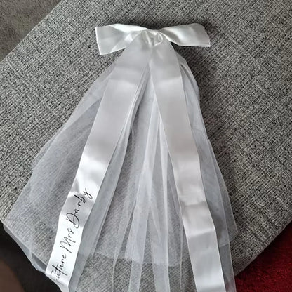 Personalized 'Future Mrs' bow veil in white tulle with satin bow, shown on bachelorette bride accessory display.